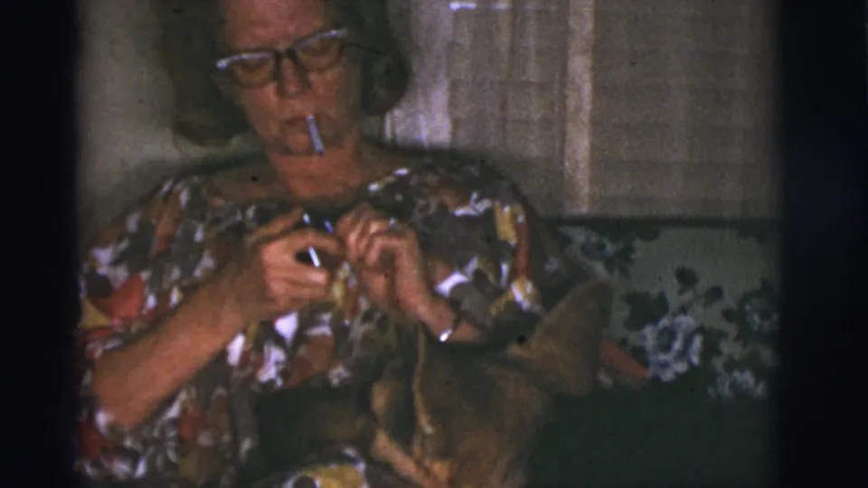 Adult woman in a floral dress and glasses lights a cigarette on a couch beside her German Shepherd in 1966 Tucson home-movie footage.
