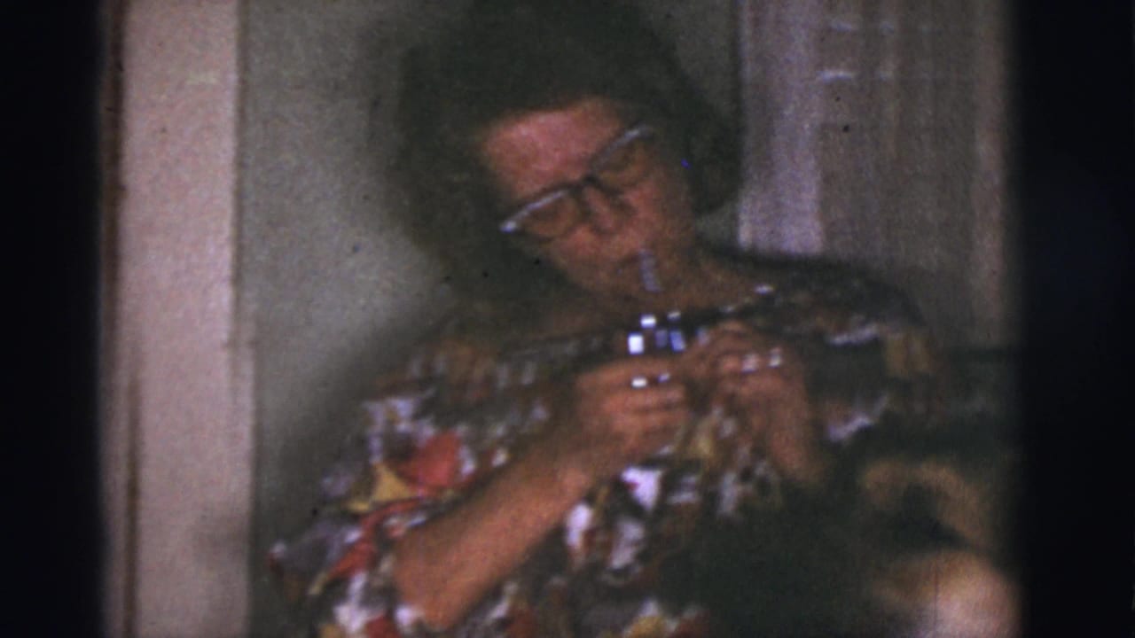 Woman in glasses and floral dress holds a cigarette and lighter in a dim interior.