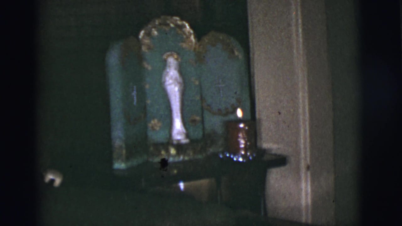 Dim home altar with Virgin Mary statue, green triptych, small crosses, and a lit candle.