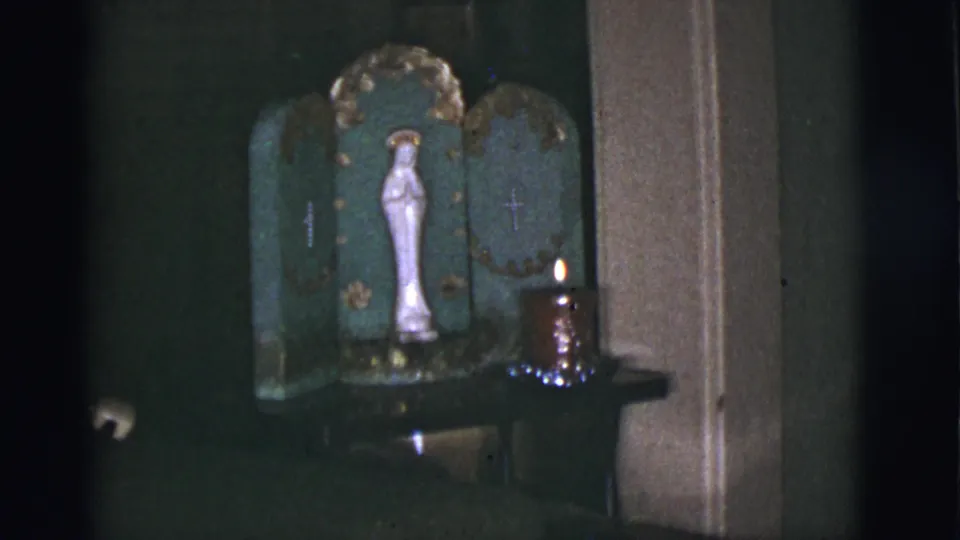 A handheld 8mm frame holds on the Catholic home altar, with the Virgin Mary statue and candle still visible.