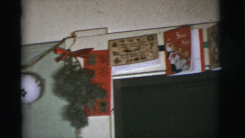 1966 Tucson-area 8mm frame showing a red holiday card, a greenery decoration, and printed Christmas cards near the end of the wall display.