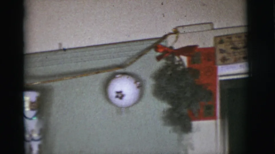 1966 Arizona home-movie frame showing a close wall display with a white star ornament, red ribbon, greenery and partial framed artwork.