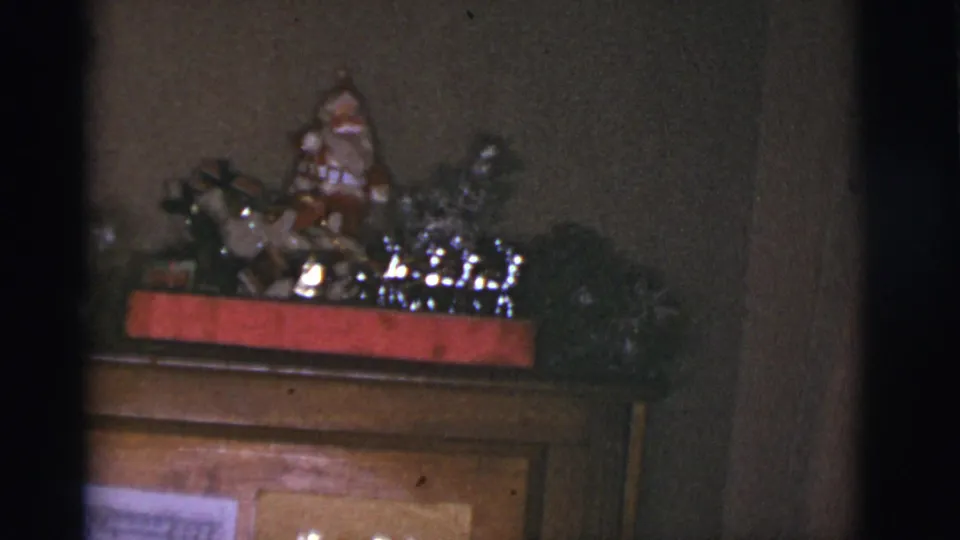 1966 Tuscon home-movie frame with a small Santa figure, greenery, and shiny decorations on a wooden cabinet.