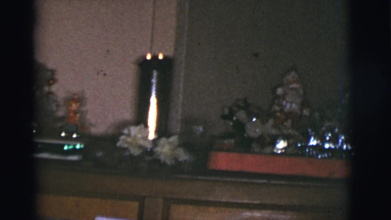 1966 Tucson home-movie frame of a holiday mantel with small figurines, shiny decorations, and a lit candle holder.