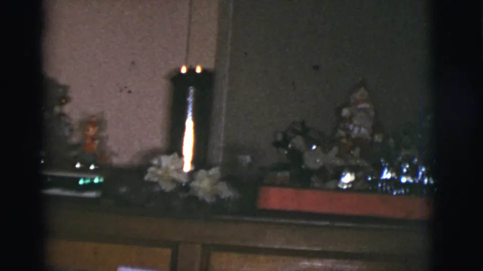 1966 Tucson home-movie frame of a holiday mantel with small figurines, shiny decorations, and a lit candle holder.