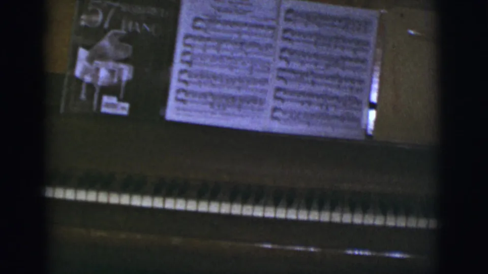 A 1966 8mm frame shows piano keys, open sheet music, and a visible book cover during a Tucson Christmas home-movie interior shot.