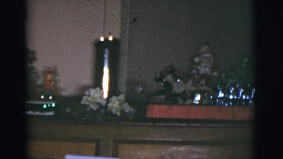 1966 Arizona home-movie frame of a piano top arranged with a lit candle, white flowers, figurines, and reflective Christmas decorations.