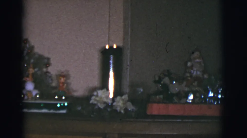 1966 Arizona home-movie frame showing small holiday figurines and decorations arranged across a piano top in a dim private room.