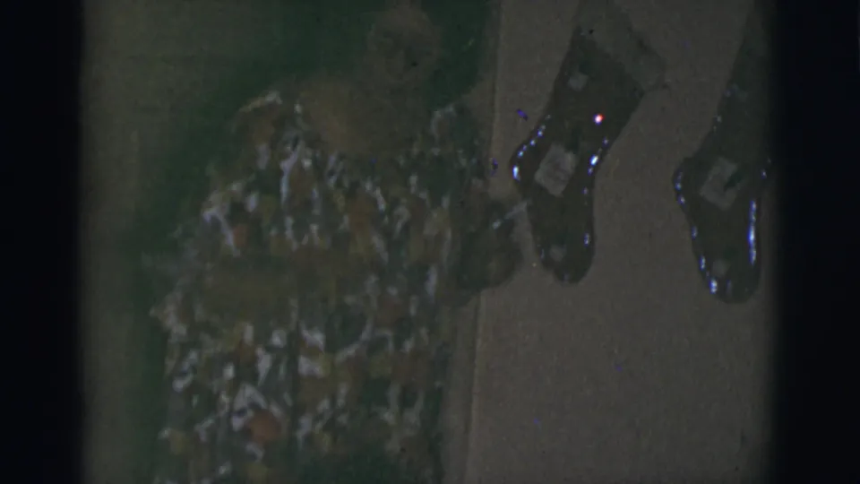 1966 Tuscon, Arizona home-movie frame with a woman in a floral dress bending toward a small dog near hanging stockings.