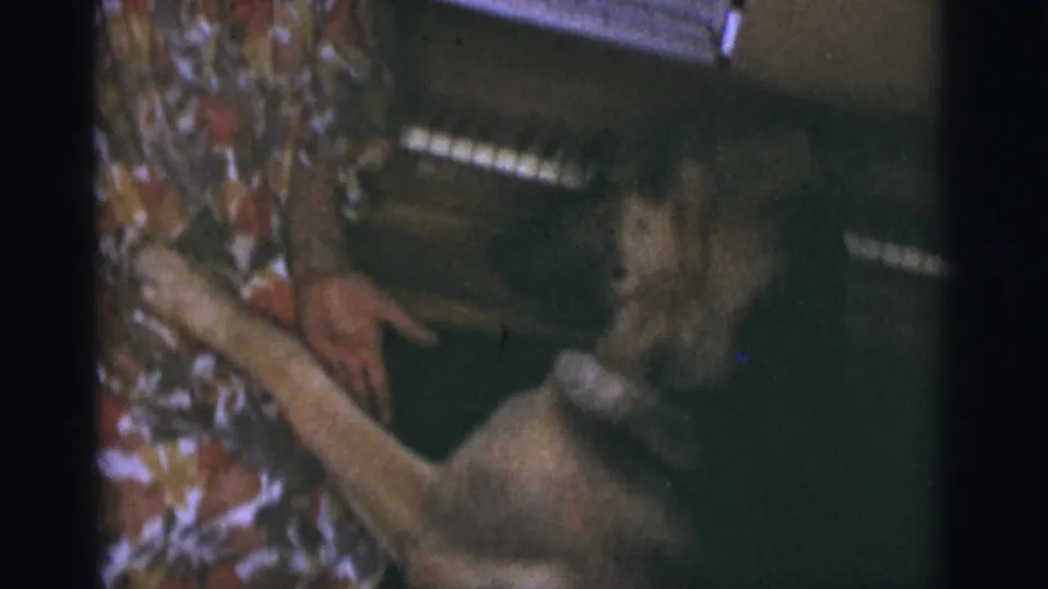 1966 Tuscon, Arizona 8mm frame with a woman reaching toward a small light-colored dog in a dim private room.