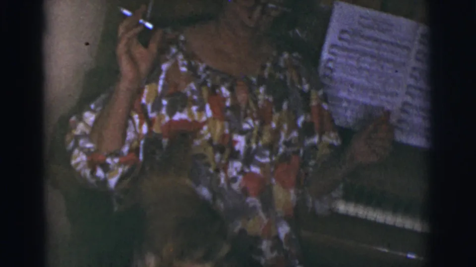 A woman in a colorful floral dress sits at the piano with sheet music visible in 1966 Tuscon home footage.