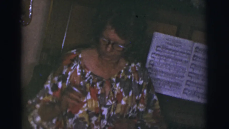 The seated woman looks down at the piano while sheet music rests on the stand in a 1960s home interior.