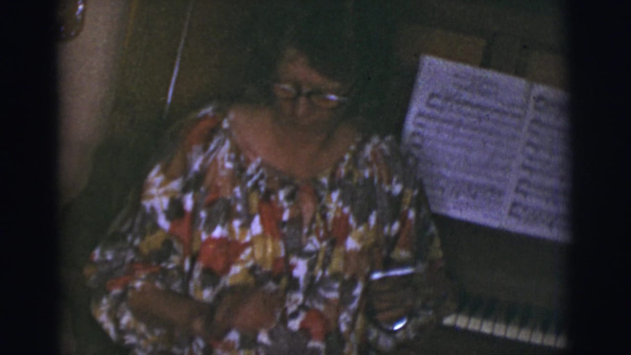 In a Tucson 1966 home interior, a woman in a colorful floral blouse sits at a piano with sheet music nearby.