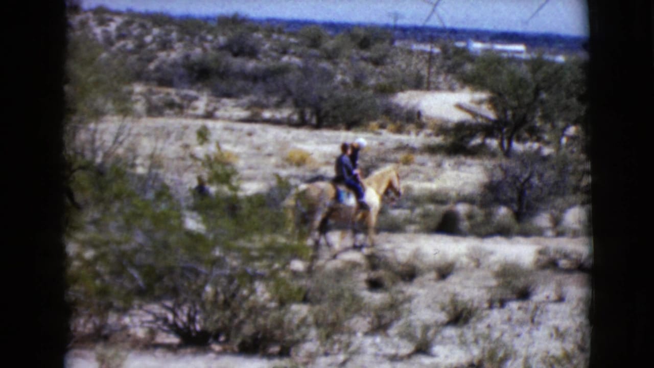 A rider on a light-colored horse crosses a desert trail with another rider behind.