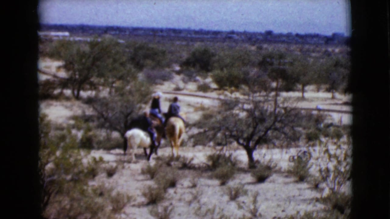 1968 Arizona home-movie frame showing two distant riders on horseback crossing sparse desert terrain near shrubs and low trees.