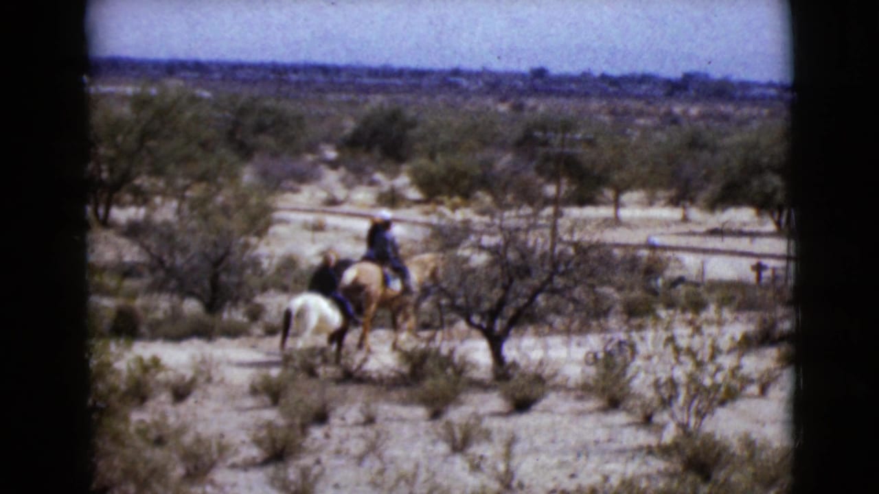 Distant riders in 1968 Arizona footage pass through arid scrubland, with a white horse and a palomino horse visible.