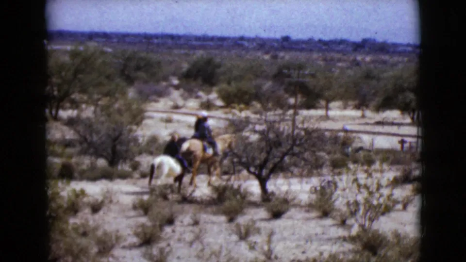 Distant riders in 1968 Arizona footage pass through arid scrubland, with a white horse and a palomino horse visible.