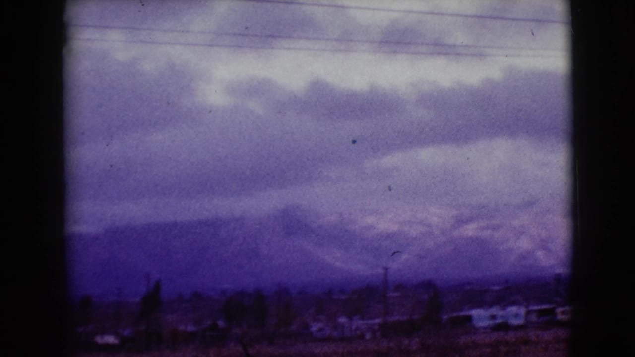 Clarksdale, Arizona 1968 home-movie frame with overcast clouds, a distant mountain range, scattered low buildings, and roadside poles.