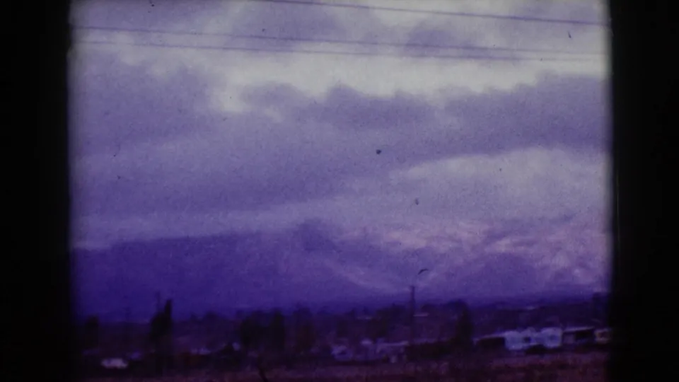 Clarksdale, Arizona 1968 home-movie frame with overcast clouds, a distant mountain range, scattered low buildings, and roadside poles.