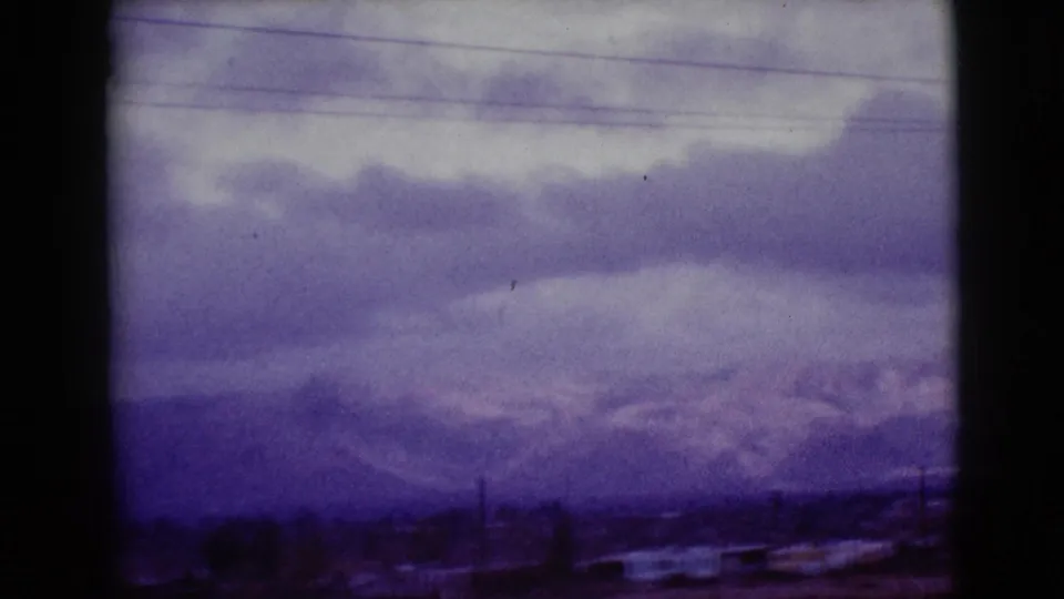 Aged 8mm frame from 1968 near Clarksdale, Arizona, with snowy mountains, layered clouds, utility wires, and a dark roadside foreground.