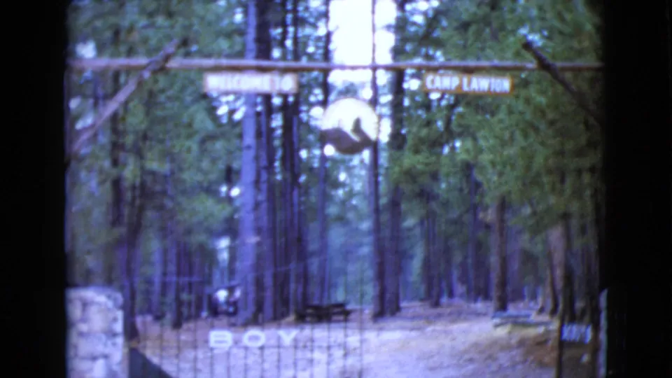 1968 8mm frame of the Camp Lawton entrance in Clarksdale, Arizona, with a wooden sign, gate bars, pine trees, and a stone pillar.
