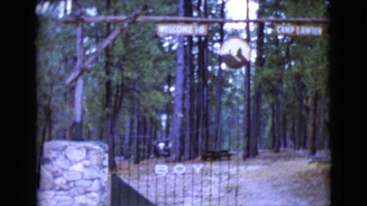 1968 8mm view of the Camp Lawton gate in Clarksdale, Arizona, showing a rustic sign, tall pines, gate bars, and a stone entry pillar.