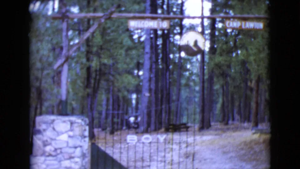 1968 8mm view of the Camp Lawton gate in Clarksdale, Arizona, showing a rustic sign, tall pines, gate bars, and a stone entry pillar.