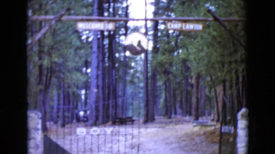 1968 8mm frame from Clarksdale, Arizona, with Camp Lawton signage, gate bars, a stone post, pine trunks, and large painted ground letters.