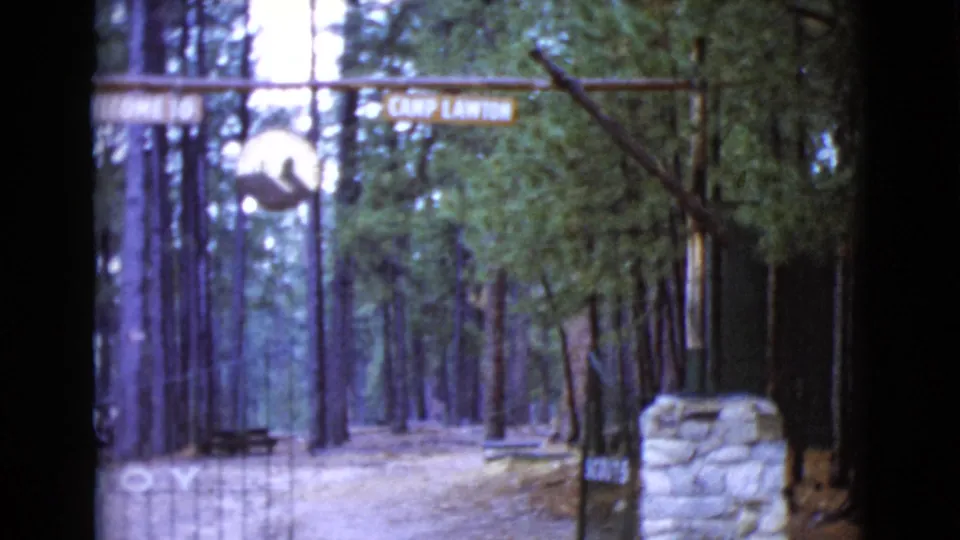 1968 8mm view of the Camp Lawton entrance in Clarksdale, Arizona, with a wooden overhead sign, pine forest, gate bars, and stone pillar.