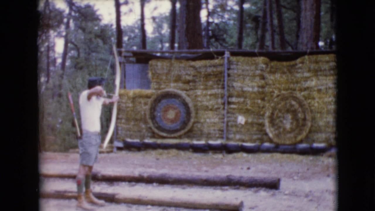 Young man draws a bow toward a straw-bale archery target in a wooded range.