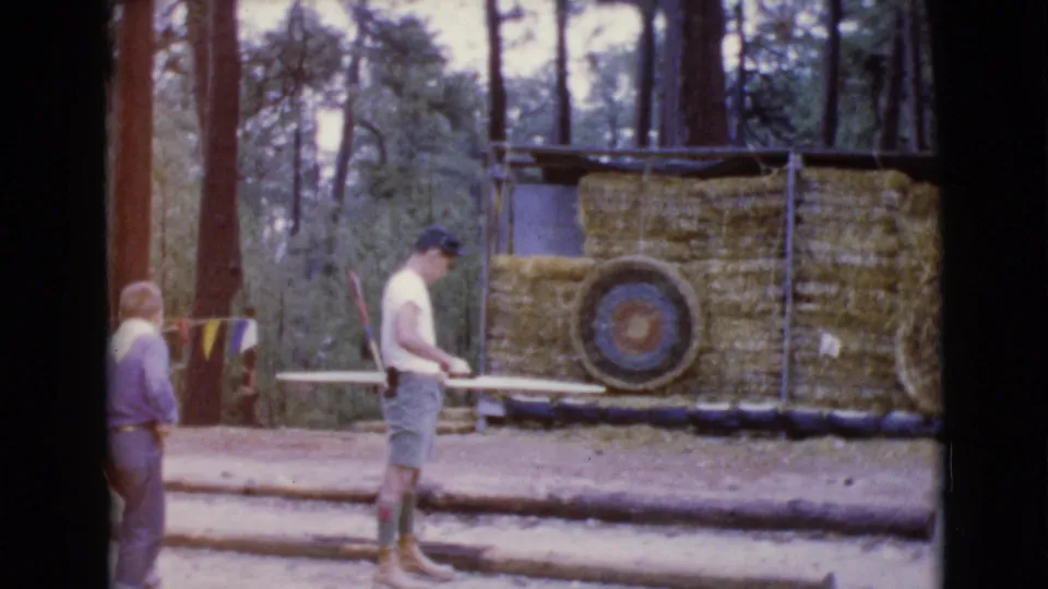 A young man examines or adjusts his bow while a second person in a purple shirt walks into the wooded range frame.