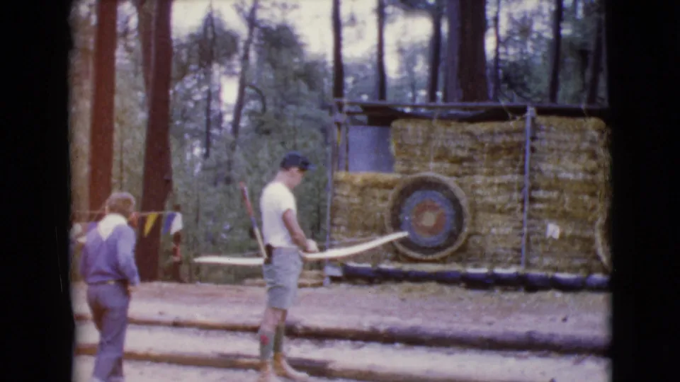 The archer stands near the straw-bale target with his bow as another person passes through the 1968 wooded range scene.