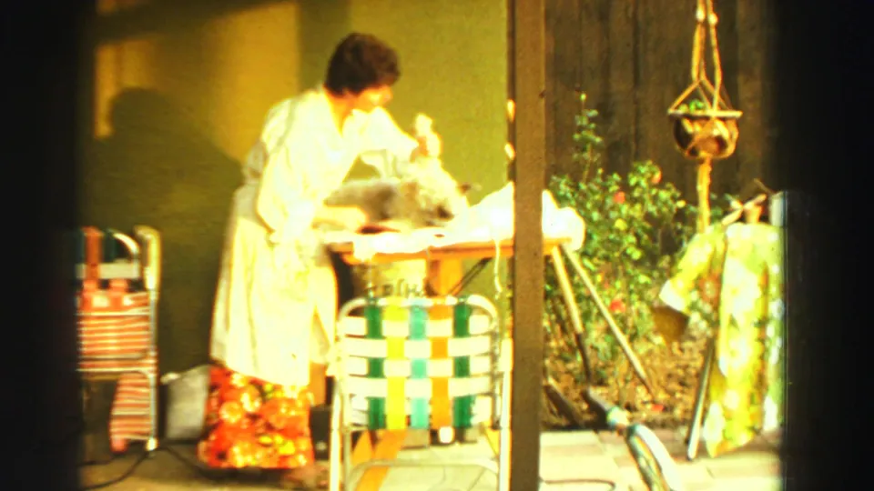 In 1969 Reno home-movie footage, a woman trims a fluffy dog on a sunlit backyard table near a woven chair and hanging planter.