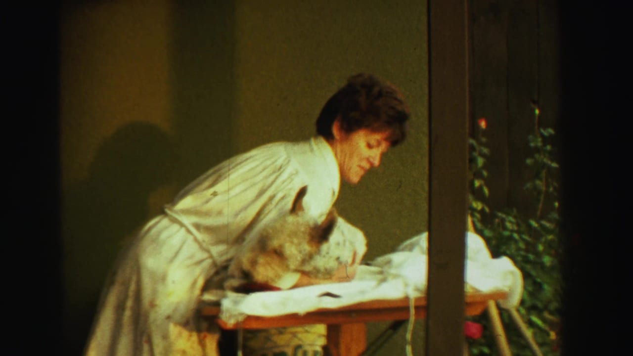 In this 1969 Reno home-movie frame, a woman in a white robe bends toward a small dog resting on a table beside greenery.