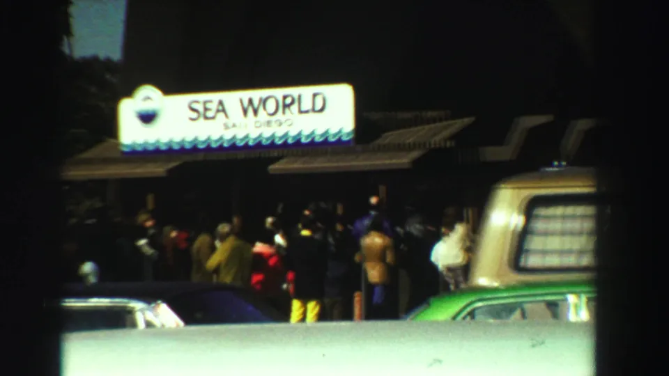 1969 home-movie frame showing visitors at the Sea World San Diego gate, with cars and a van partly blocking the lower view.