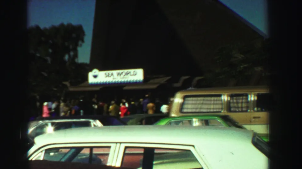 Wider 1969 8mm view of the Sea World San Diego entrance, showing a visitor crowd behind parked cars and a van.