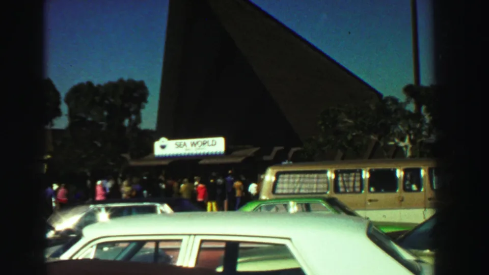 1969 Reno home-movie frame showing the Sea World entrance, visitor crowd, classic cars, tan van, and steep pointed-roof structure.