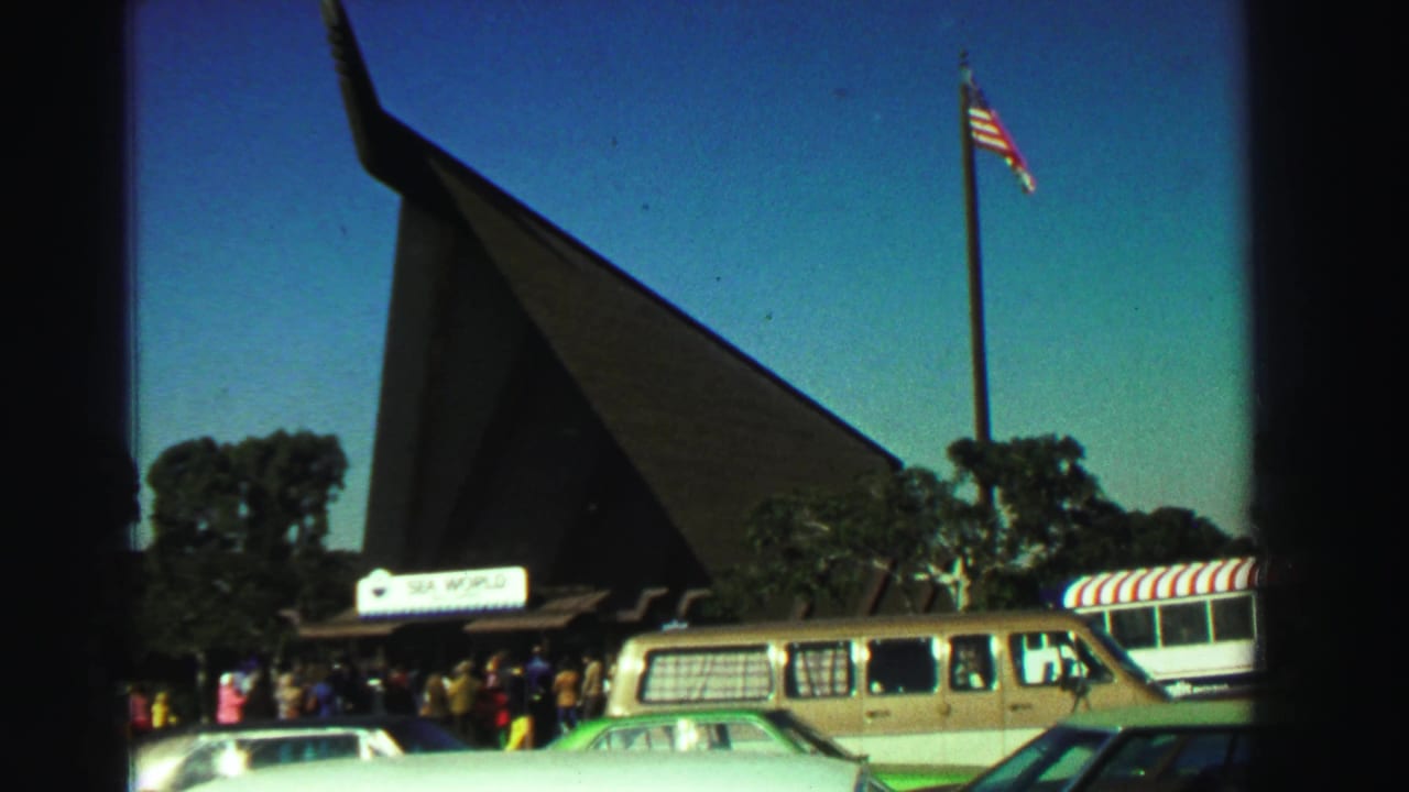 1969 Reno frame showing the Sea World entrance from the parking area, with classic cars, a tan van, visitors, trees, and a waving flag.