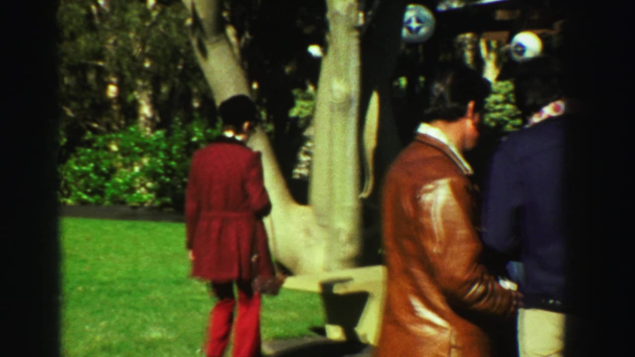 1969 Reno park visitors seen from behind and in profile, including a person in red and another in a brown leather jacket near trees and benches.