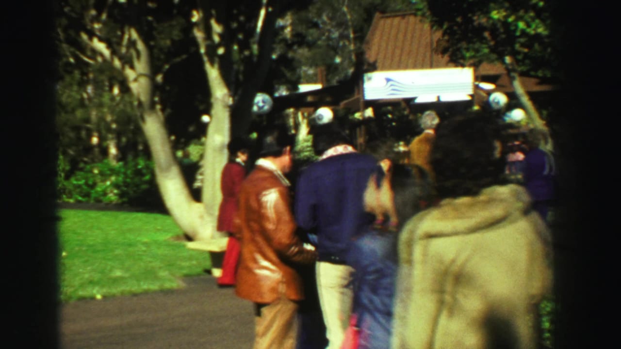 1969 Reno park path with several visitors walking or standing, surrounded by trees, benches, lawn, and a small structure or sign area.