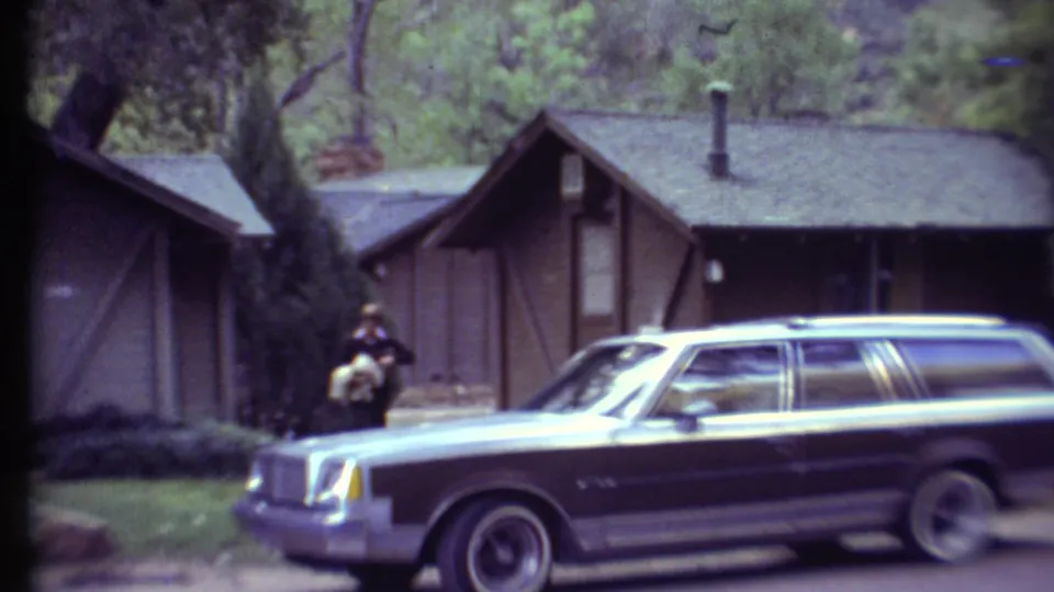 1972 Grand Canyon campground scene with a station wagon parked in front of cabins and a man visible near a large tree.