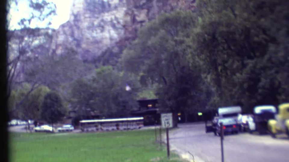 1972 Grand Canyon campground road with parked cars, trees, small signs, and canyon-like rock formations visible beyond the wooded area.