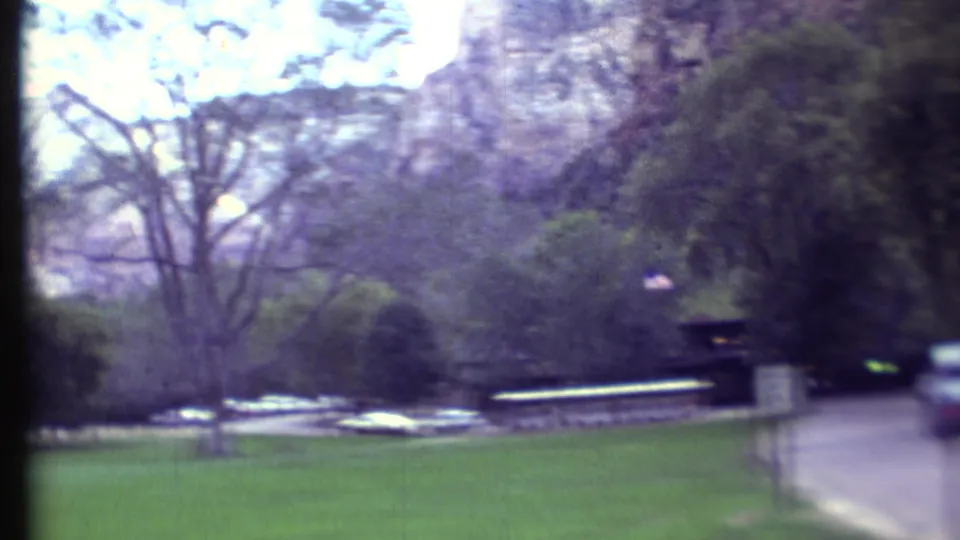 Soft 8mm frame of a quiet park road, green grounds, mature trees, parked cars, and pale cliffs in the background.