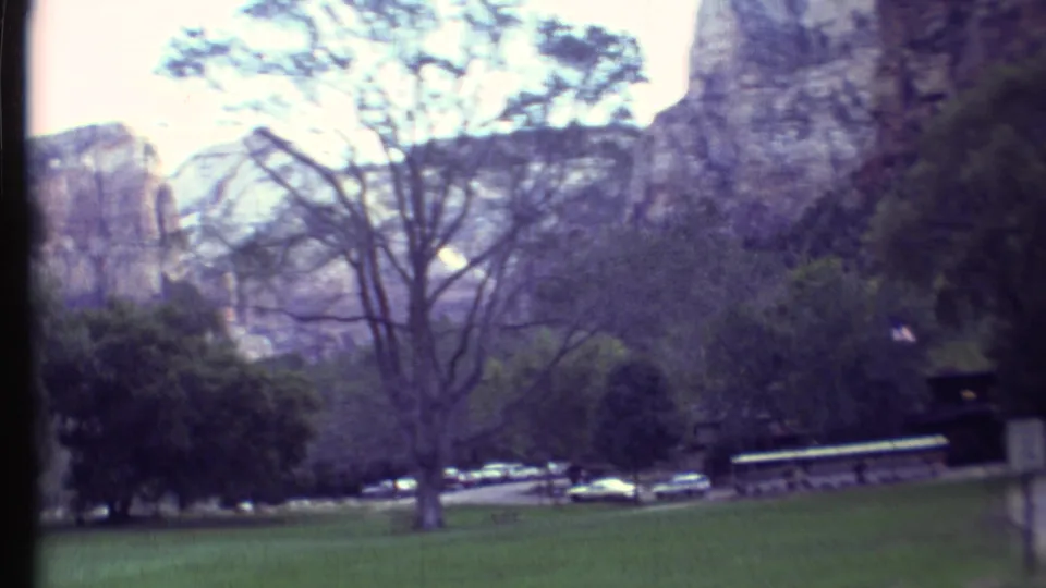 Visitor-shot 1970s frame across open grass, trees, roadside parking, a small sign, and cliff formations in the supplied park setting.