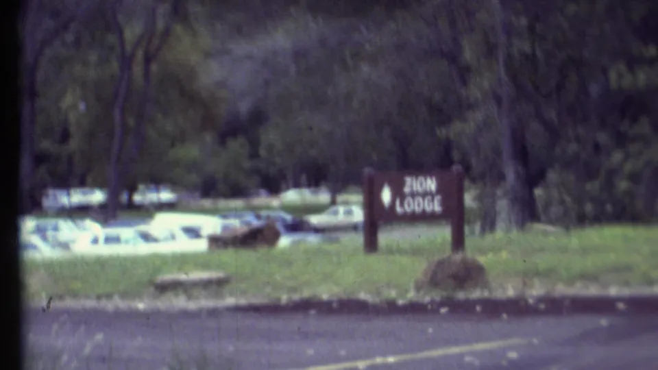 1972 8mm frame showing a Zion Lodge sign beside grassy grounds, trees, parked cars, and a paved road.
