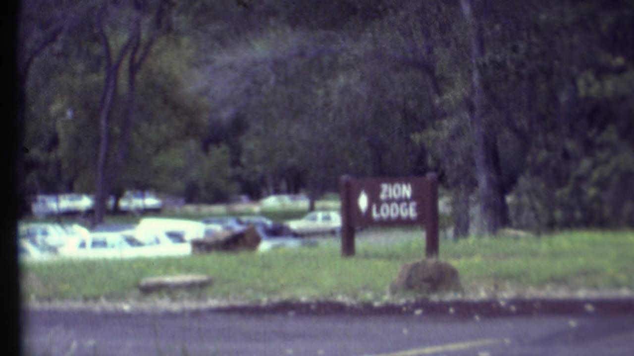 Zion Lodge sign beside a road with parked cars and trees behind it.