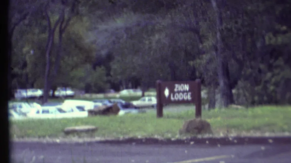 1972 8mm travel frame showing a Zion Lodge sign on a grassy verge, with parked cars, trees, and a paved road in view.