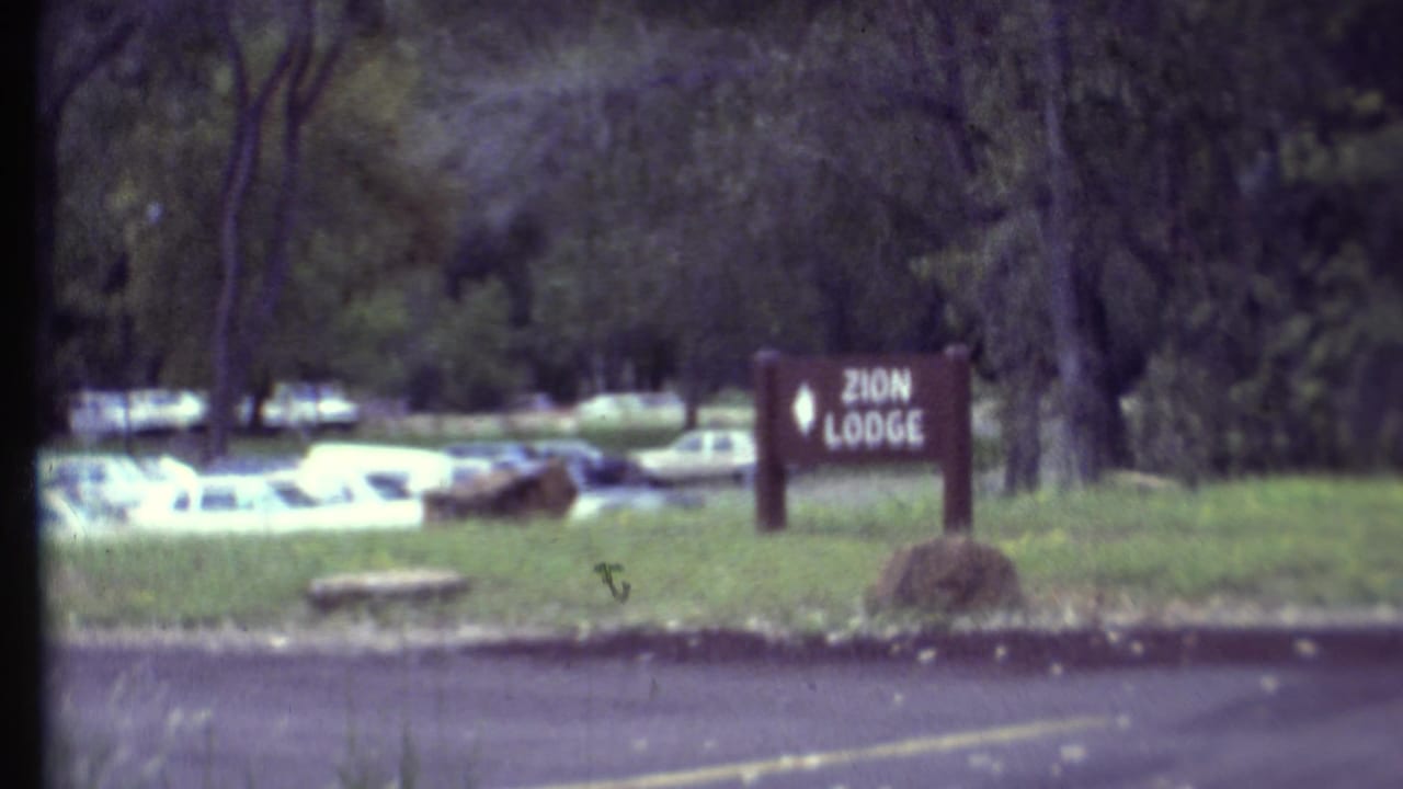 Zion Lodge sign near trees and parked cars across a paved road.