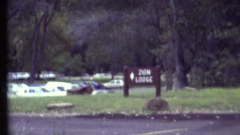 1972 8mm travel frame showing the Zion Lodge sign from the roadside, with parked cars and dense trees behind it.