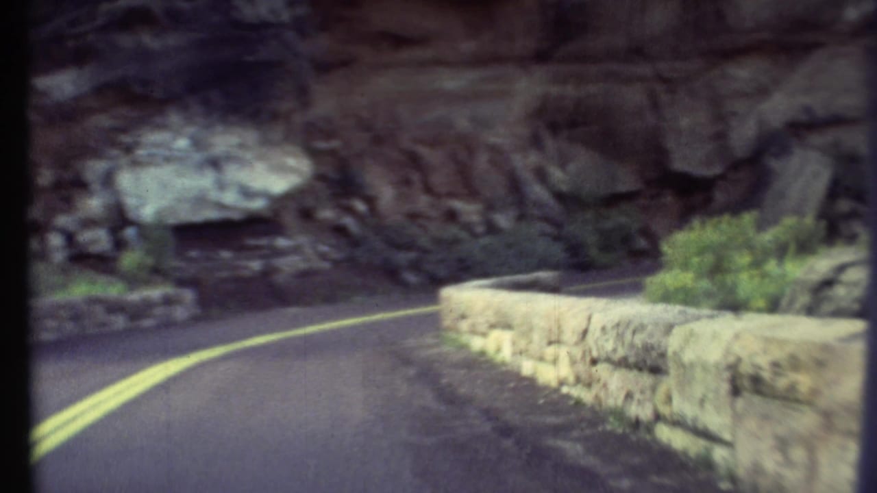 1972 8mm Grand Canyon road view showing a paved curve, double yellow lines, stone wall, rock face, and desert vegetation.